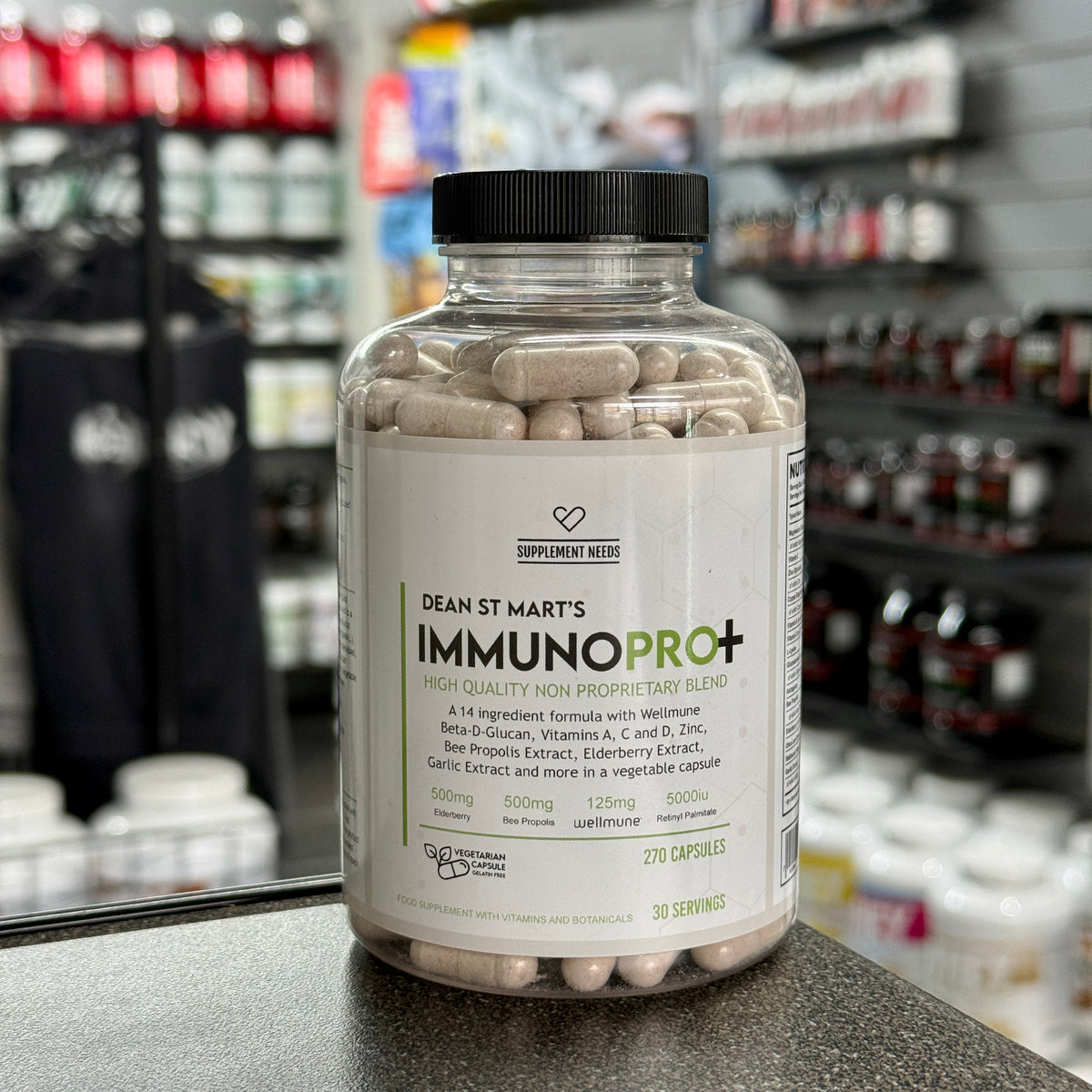 A clear supplement bottle labeled Dean St. Mart's Supplement Needs ImmunoPro+ with a black cap, filled with beige capsules, sitting on a countertop in a blurred retail store setting.