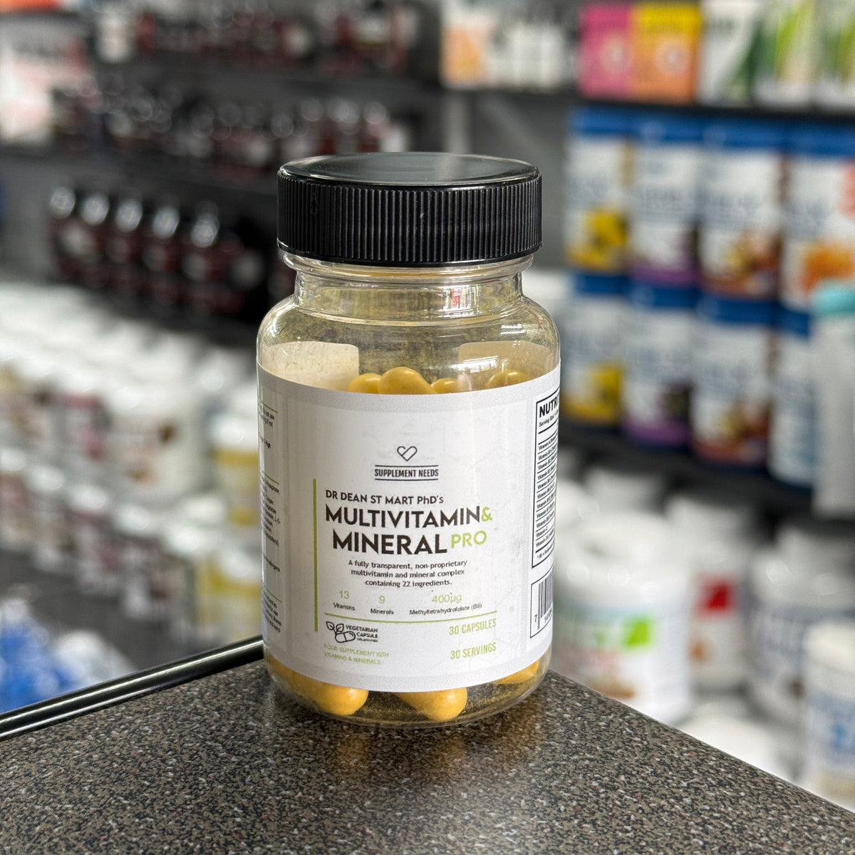 A bottle of Dr. Dean St Mart's Multivitamin & Mineral Pro dietary supplement, containing 30 capsules, placed on a dark countertop with a blurred supplement store background.