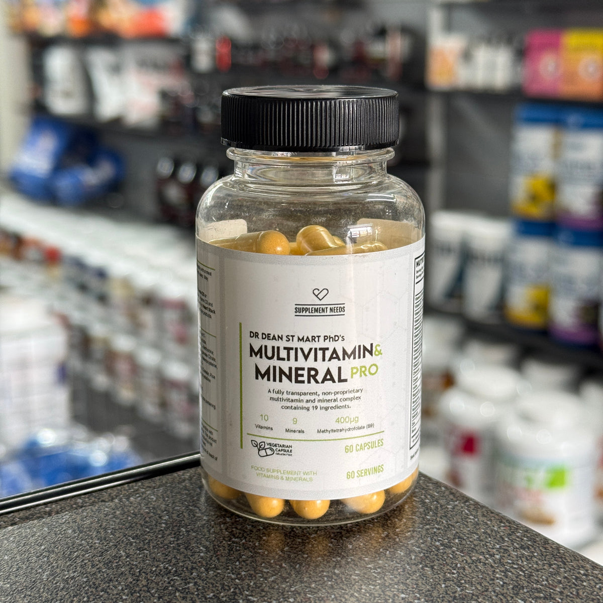 A bottle of Dr. Dean St. Marts multivitamin and mineral pro 60 capsules against a blurred supplement store background