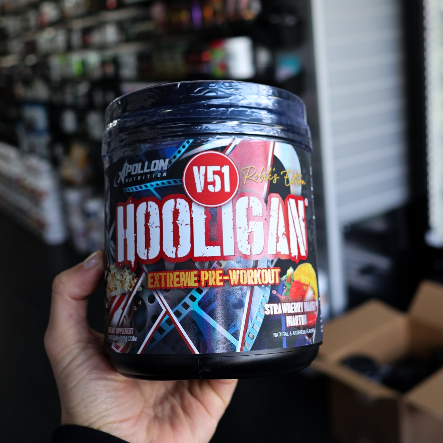 Container of V51 Hooligan pre-workout supplement held in a hand with a store shelf in the background.