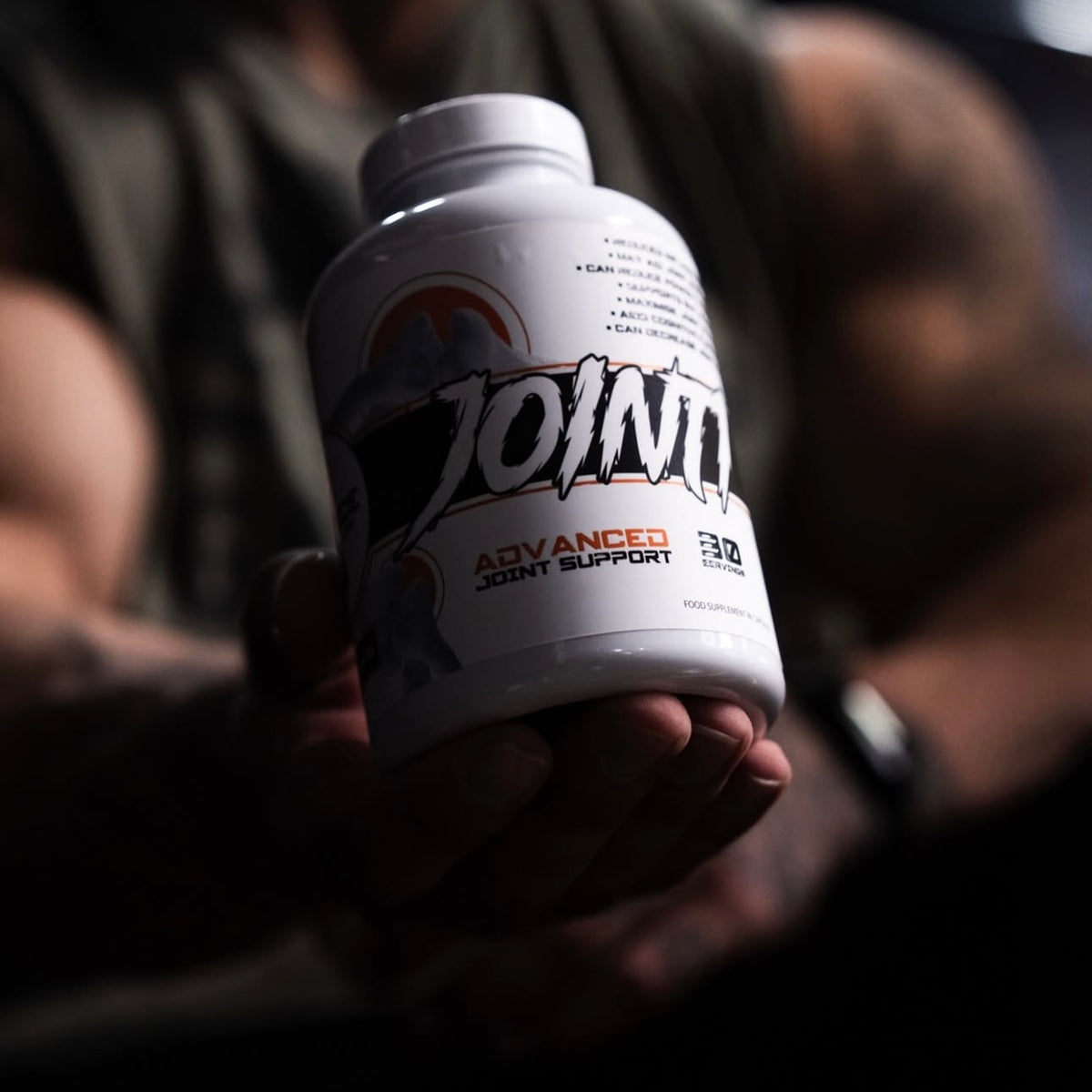 A hand holding a white bottle of Joint 1 Advanced Joint Support supplement, with a blurred background showing a person's arms.
