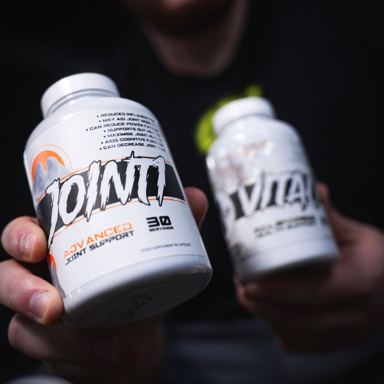 A close-up lifestyle shot of a person holding two white supplement bottles. The foreground bottle is labeled 'JOINT Advanced Joint Support' and features a prominent black and white logo with orange accents.