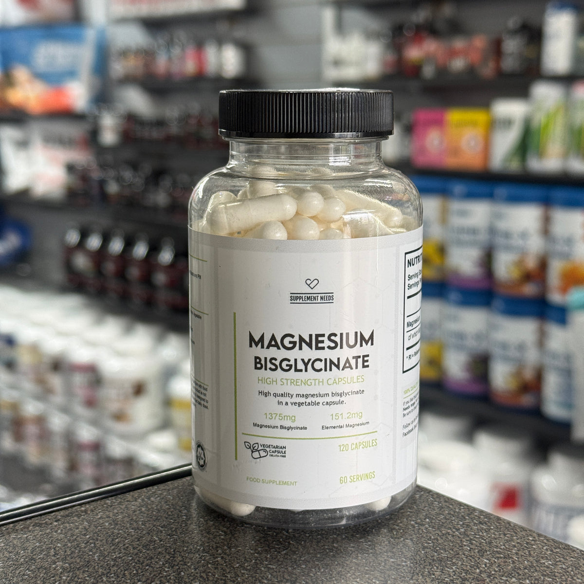 A bottle of Dr. Dean St Mart's Supplement Needs Magnesium Bisglycinate dietary supplement capsules, containing 120 capsules, set on a countertop with a blurred pharmacy or supplement store background.