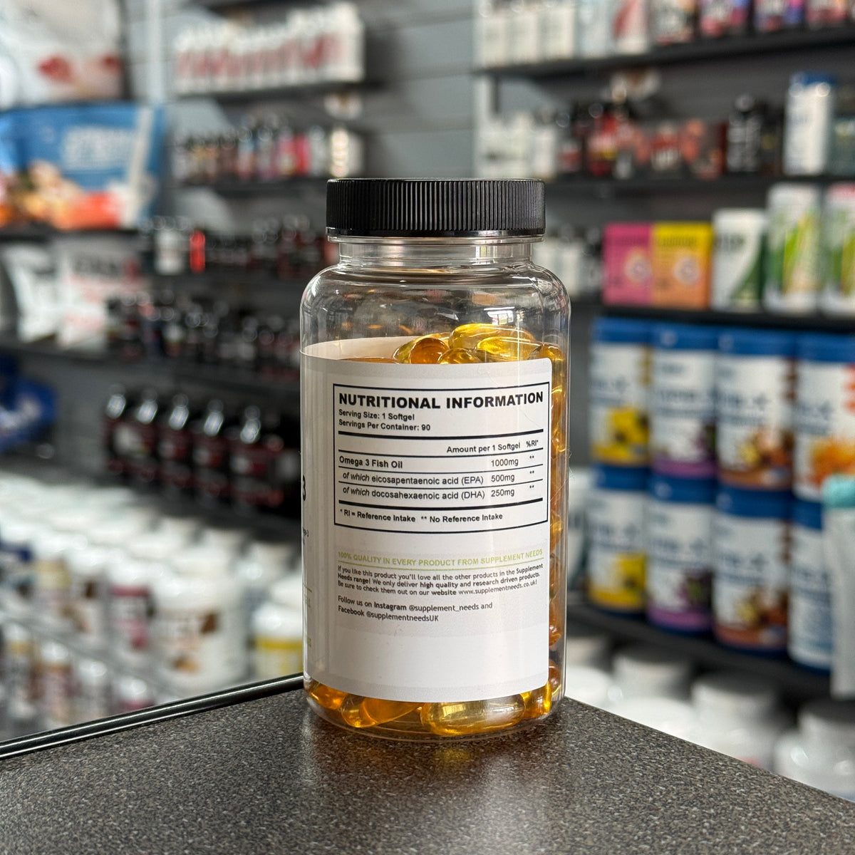 A clear plastic supplement bottle filled with yellow softgels, featuring a black ribbed cap and a white label with nutritional information, set on a countertop with a blurred supplement store background.