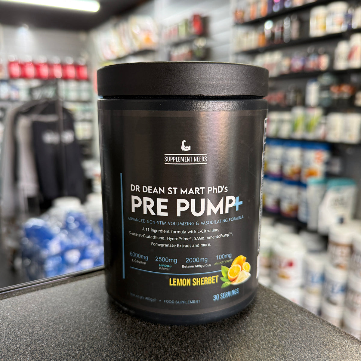 A black tub of Supplement Needs Pre Pump+ in Lemon Sherbet flavour, with white and blue text, highlighting the brand, product name, and contents (30 servings), on a blurred shop background.